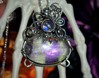 May include: A silver pendant necklace featuring a large, oval-shaped gemstone with purple and green hues. The stone is encased in a decorative silver wire setting with a smaller, round, iridescent stone at the top. The necklace is displayed on a white skeleton hand.