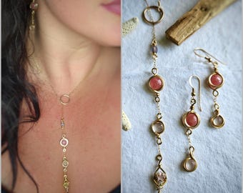 May include: A gold-toned lariat necklace and matching drop earrings featuring small circular frames. The jewelry set incorporates pink and clear round gemstones, along with delicate gold-colored metal chains and wire-wrapped accents.