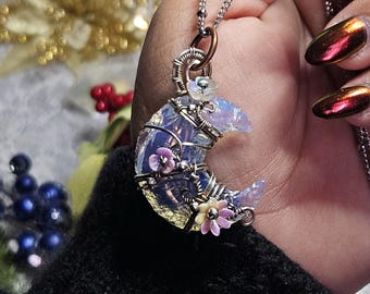 May include: A handcrafted crescent moon pendant necklace. The pendant features a wire-wrapped, iridescent, blue and purple glass moon adorned with small pink and white flowers. The necklace has a silver chain.