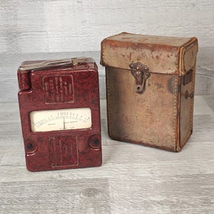 May include: A maroon electrical testing device with a gauge and a silver handle, alongside a worn brown leather case with a metal clasp. The device has text on its face.