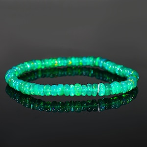Puede incluir: Un brazalete de ópalo verde. El brazalete está compuesto por pequeñas cuentas de ópalo redondas y pulidas. Las cuentas tienen una mezcla de tonos verdes y amarillos, con una superficie reflectante. El brazalete se muestra sobre una superficie oscura y reflectante.