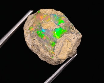 May include: A rough, uncut opal gemstone held by metal tweezers. The opal displays vibrant flashes of green, orange, and blue within a matrix of tan-colored rock. The gemstone is set against a black background.