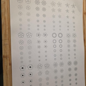May include: A white paper with black line drawings of various flower templates. The text "Groombridge Opals Templates" is at the top, and "Jewellery Template Flowers 6mm - 15mm" is at the bottom. The paper is on a wooden surface.