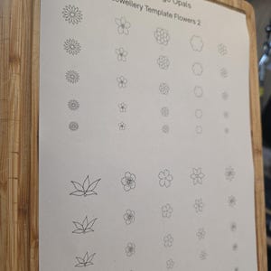 May include: A sheet of paper with various small flower designs, labeled "Groombridge Opals Jewellery Template Flowers 2." The flowers are in different styles and sizes, all drawn in black ink. The paper is on a wooden cutting board.