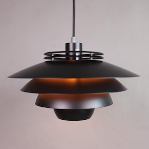 Peut inclure: Un luminaire suspendu noir moderne avec un design en gradins en forme de soucoupe. Le luminaire a une finition mate et une lueur chaude émanant de l'intérieur. La lumière est suspendue à un cordon noir.