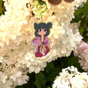 Peut inclure: Un porte-clés en acrylique transparent représentant un personnage de dessin animé aux cheveux bleu foncé coiffés en deux chignons, portant une robe traditionnelle rose et magenta avec une ceinture jaune, fixé à un mousqueton en plastique vert lime.