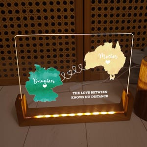 May include: A lighted acrylic sign with a wooden base. The sign features a map of Germany and Australia, labeled "Daughter" and "Mother," connected by a dotted line of hearts. The text "THE LOVE BETWEEN KNOWS NO DISTANCE" is below the maps.