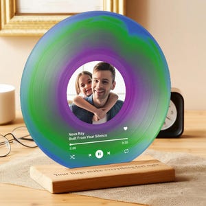 以下が含まれることがあります： 男性と子供の写真が入った、レコード盤型のパーソナライズされたフォトフレーム。レコードは青、緑、紫のグラデーション。木製のベースには「Your hugs make everything feel right」の文字。
