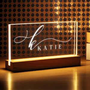 Può includere: Una luce notturna a LED personalizzata con il nome "Katie" in corsivo. Il pannello in acrilico trasparente è illuminato da una luce bianca calda, posto su una base rettangolare in legno. Lo sfondo presenta un motivo intrecciato.
