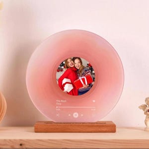 Könnte beinhalten: Ein rosa, kreisförmiger Fotorahmen, der einer Schallplatte ähnelt. In der Mitte befindet sich ein Foto von zwei lächelnden Personen, die Geschenke halten. Der Rahmen steht auf einem Holzsockel. Der Text "The Best Tina" und eine Musikplayer-Grafik sind sichtbar.