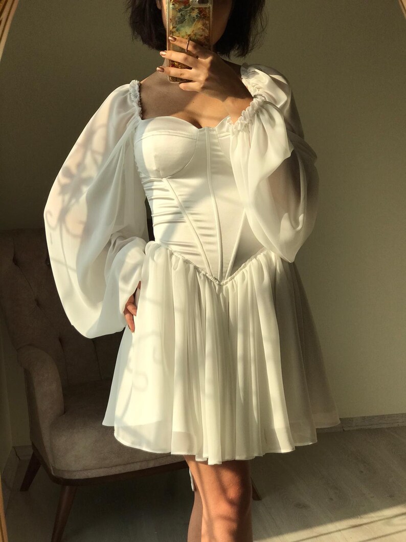 May include: A white mini dress with a sweetheart neckline and long, puffy sleeves. The dress has a fitted bodice and a flowing skirt.