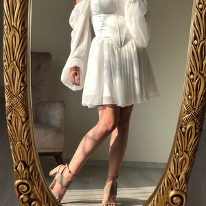 May include: A white off-the-shoulder dress with a corset bodice and long, puffy sleeves. The dress has a short, A-line skirt and is made of a lightweight, flowing fabric.