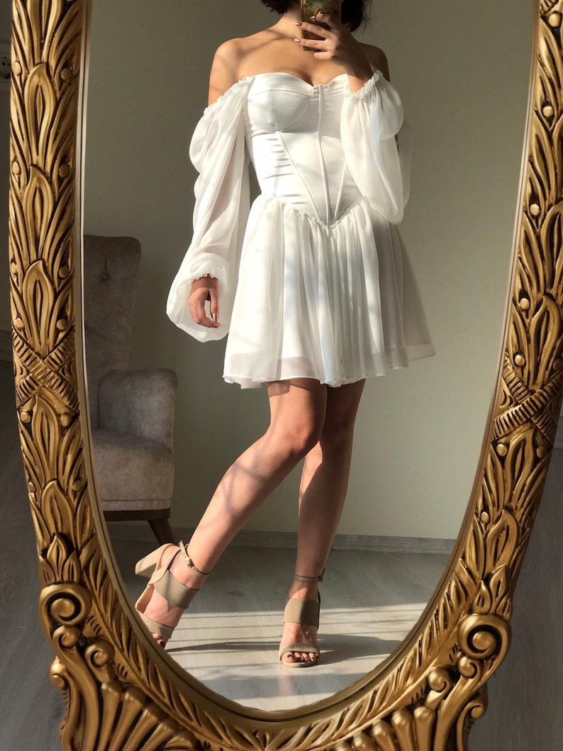 May include: A white off-the-shoulder dress with a corset bodice and long, puffy sleeves. The dress has a short, A-line skirt and is made of a lightweight, flowing fabric.