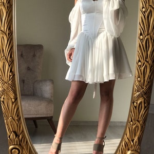 May include: A white off-the-shoulder dress with a corset bodice and puffy sleeves. The dress has a short, flowing skirt and is reflected in a gold-framed mirror.
