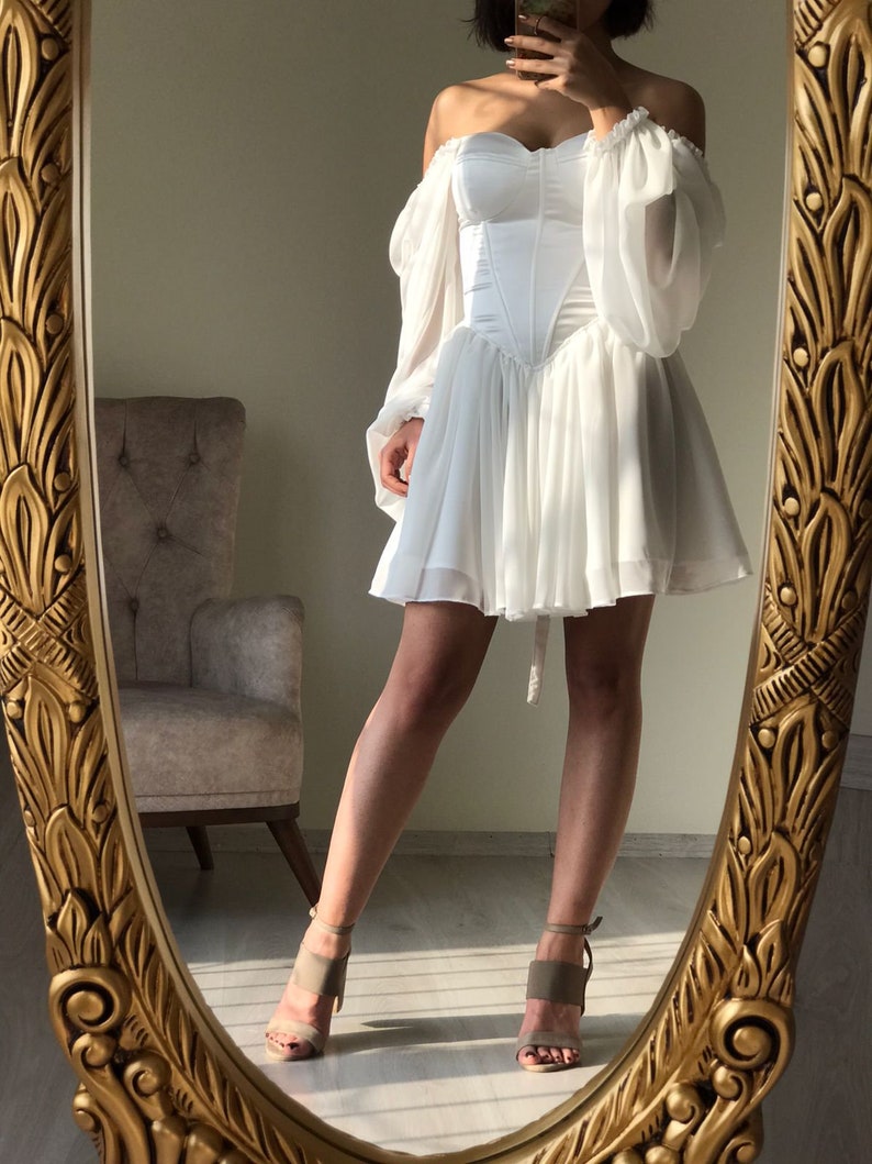 May include: A white off-the-shoulder dress with a corset bodice and puffy sleeves. The dress has a short, flowing skirt and is reflected in a gold-framed mirror.