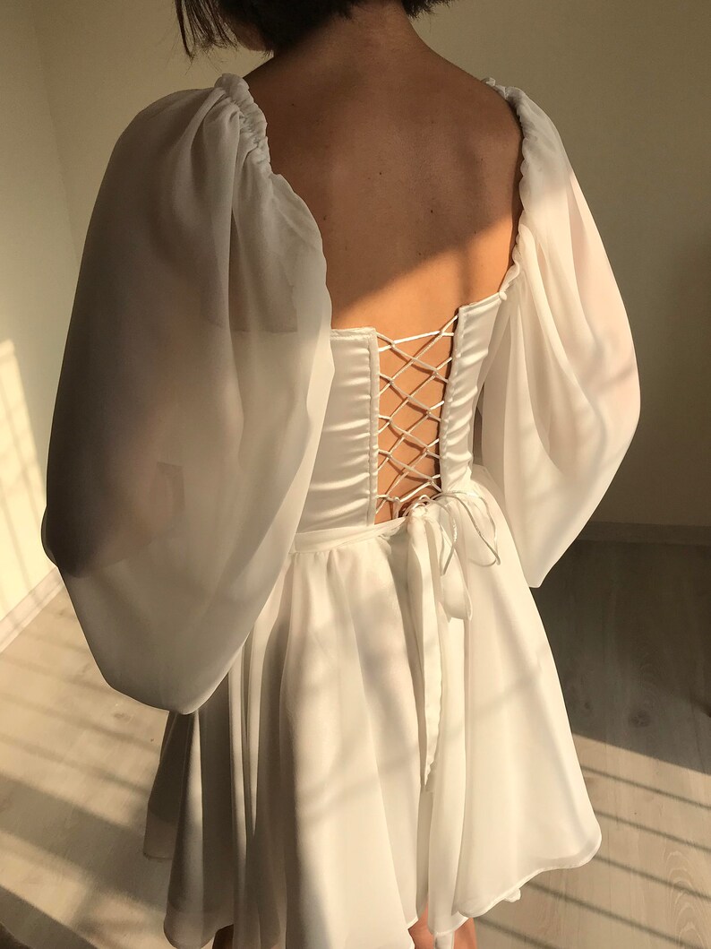 May include: White dress with a lace-up back and puff sleeves. The dress has a square neckline and a flowy skirt.