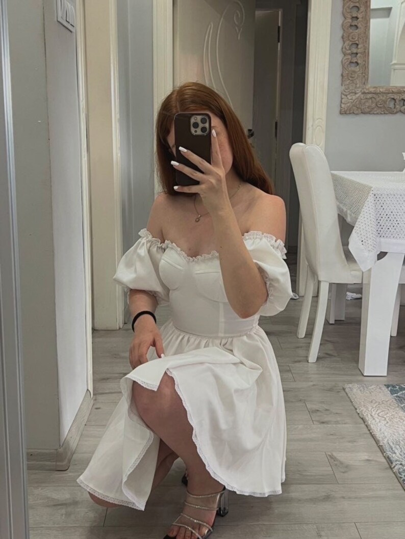 May include: A white off-the-shoulder dress with puffed sleeves and a lace trim. The dress has a fitted bodice and a flowing skirt.