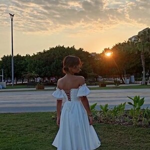 May include: A woman wearing a white off-the-shoulder dress with a lace-up back. The dress has a fitted bodice and a full skirt. She is standing in a park with green grass and trees in the background. The sun is setting in the distance.
