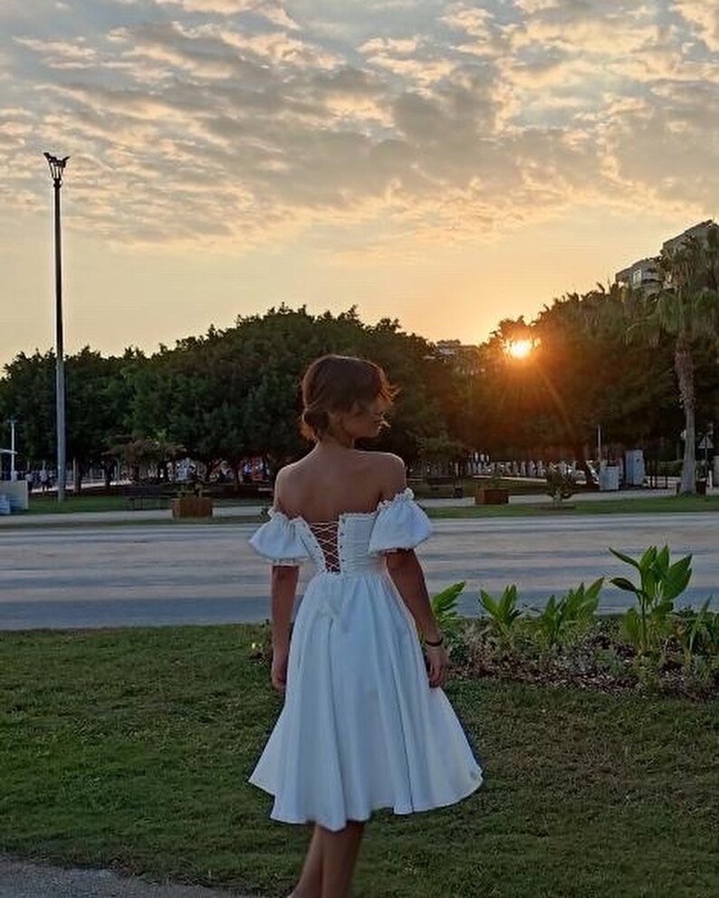 May include: A woman wearing a white off-the-shoulder dress with a lace-up back. The dress has a fitted bodice and a full skirt. She is standing in a park with green grass and trees in the background. The sun is setting in the distance.