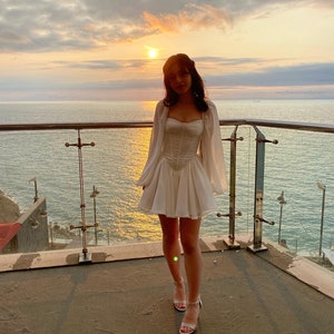 May include: A woman wearing a white dress with a ruffled skirt and long sleeves stands on a balcony overlooking the ocean. The sun is setting in the background, casting a warm glow on the scene.