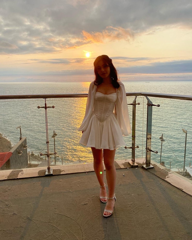 May include: A woman wearing a white dress with a ruffled skirt and long sleeves stands on a balcony overlooking the ocean. The sun is setting in the background, casting a warm glow on the scene.