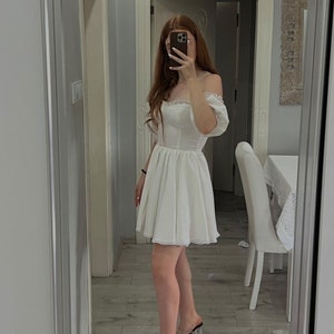 May include: A white off-the-shoulder dress with a lace trim and a fitted bodice. The dress has a full skirt and is made of a lightweight fabric. The model is wearing clear heeled sandals.