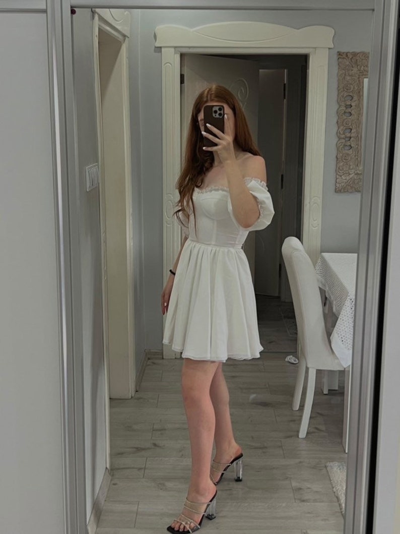 May include: A white off-the-shoulder dress with a lace trim and a fitted bodice. The dress has a full skirt and is made of a lightweight fabric. The model is wearing clear heeled sandals.