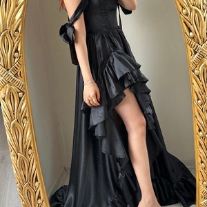 May include: A black off-the-shoulder gown with a high-low hemline and ruffled detailing. The dress features a fitted bodice and flowing skirt, with shoulder ties. The dress is reflected in a gold-framed mirror.