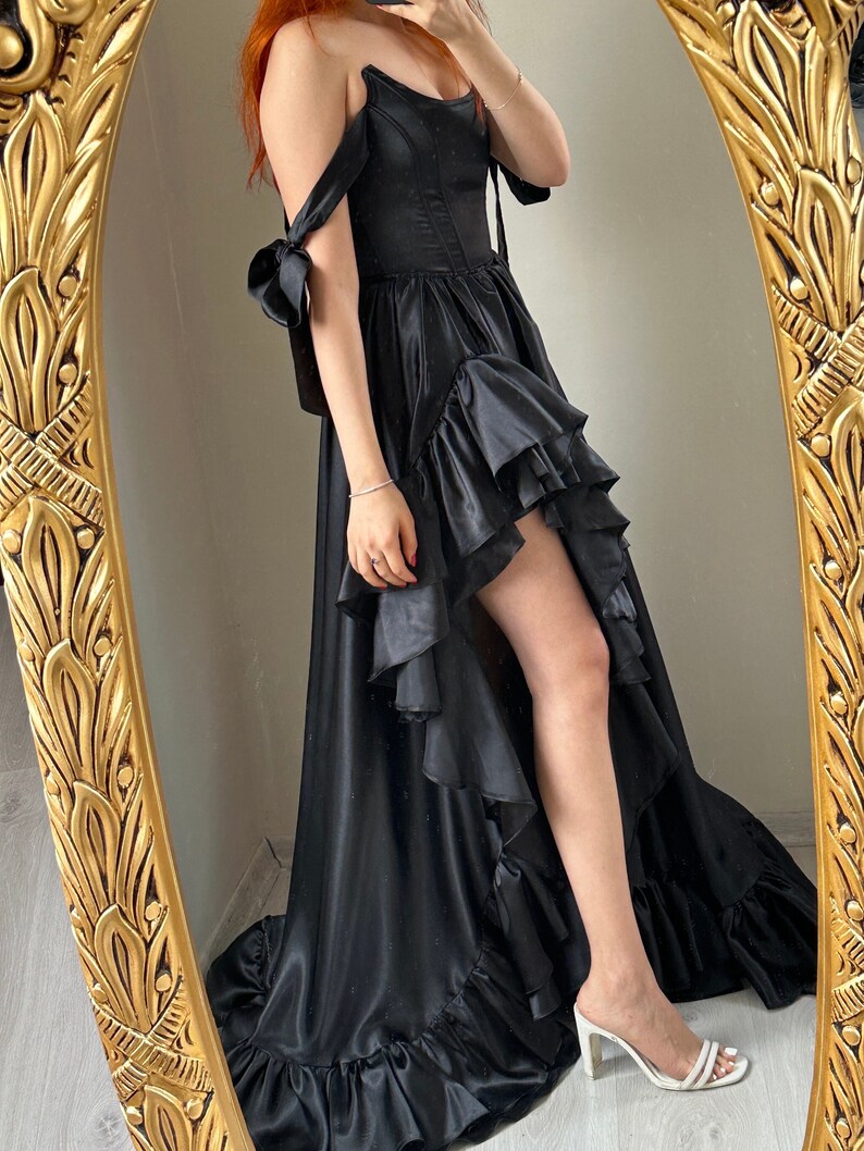 May include: A black off-the-shoulder gown with a high-low hemline and ruffled detailing. The dress features a fitted bodice and flowing skirt, with shoulder ties. The dress is reflected in a gold-framed mirror.