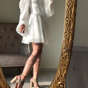 May include: A white off-the-shoulder dress with a fitted bodice and a flowing skirt. The dress has long, puffy sleeves and is cinched at the waist. The model is wearing the dress with nude heels.