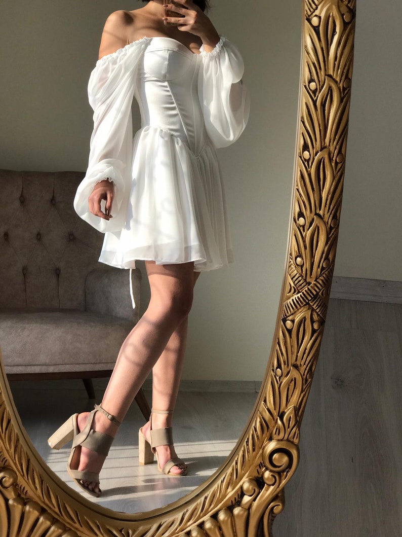 May include: A white off-the-shoulder dress with a fitted bodice and a flowing skirt. The dress has long, puffy sleeves and is cinched at the waist. The model is wearing the dress with nude heels.