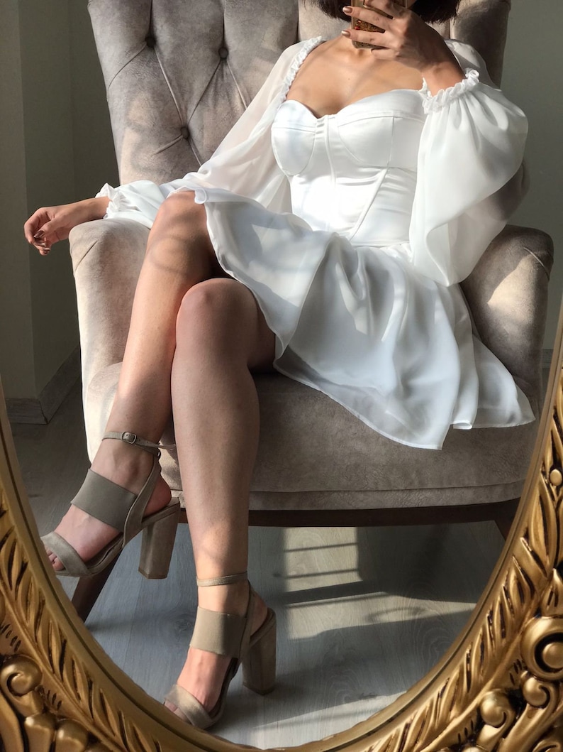 May include: A white dress with a sweetheart neckline and puffy sleeves. The dress is made of a satin-like fabric and has a short, A-line skirt. The model is wearing the dress with nude heels.