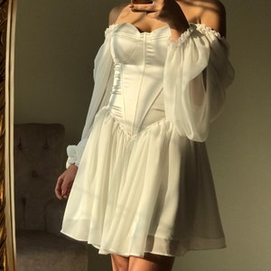 May include: A white off-the-shoulder dress with a fitted bodice and a flowing skirt. The dress has long sleeves with puffed shoulders and a sweetheart neckline.
