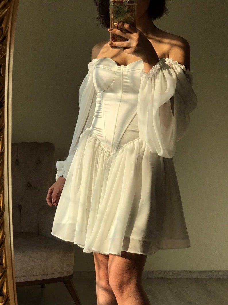 May include: A white off-the-shoulder dress with a fitted bodice and a flowing skirt. The dress has long sleeves with puffed shoulders and a sweetheart neckline.