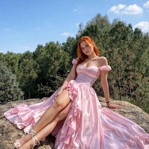 May include: A light pink satin gown with a high-low hemline and ruffled details. The off-the-shoulder bodice and skirt are a soft pink. The dress is worn outdoors on a rock with a backdrop of trees and a blue sky.