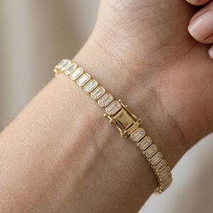 Op de afbeelding: Een gouden armband met rechthoekige, heldere edelstenen. De armband is vastgemaakt met een gouden sluiting. De edelstenen zijn in een rij gerangschikt, waardoor een doorlopende, sprankelende band ontstaat. De armband wordt om een pols gedragen.
