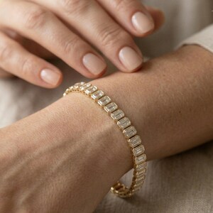 Op de afbeelding: Een gouden armband met rechthoekige, heldere stenen wordt om een pols gedragen. De armband is gemaakt van goud en heeft een reeks rechthoekig geslepen stenen die dicht bij elkaar zijn geplaatst. De huidskleur is licht, de nagels zijn neutraal gelakt.