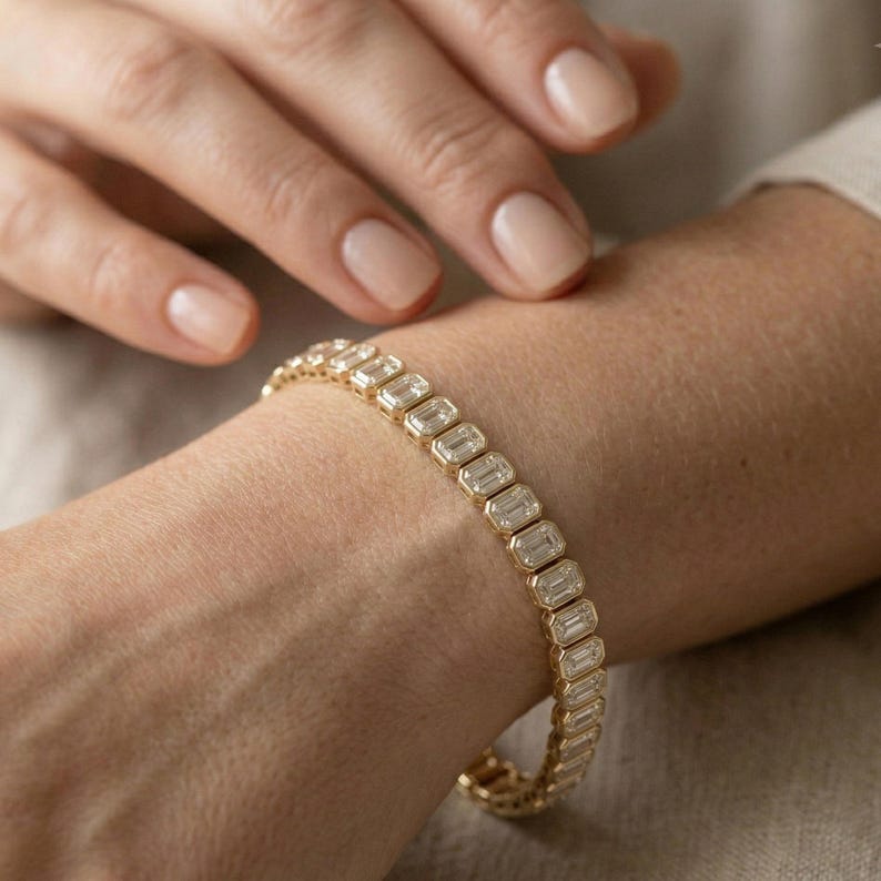 Op de afbeelding: Een gouden armband met rechthoekige, heldere stenen wordt om een pols gedragen. De armband is gemaakt van goud en heeft een reeks rechthoekig geslepen stenen die dicht bij elkaar zijn geplaatst. De huidskleur is licht, de nagels zijn neutraal gelakt.