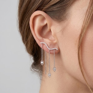 May include: Silver-toned ear cuff earring featuring a curved bar set with small, clear stones. Three fine chains dangle, each with a teardrop-shaped clear stone. The earring is worn on the ear.