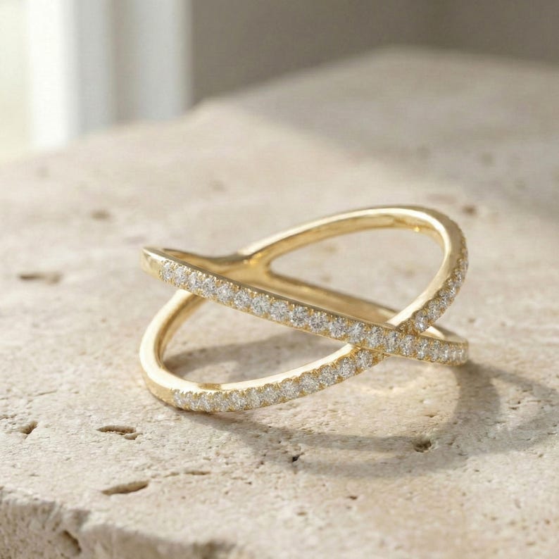 Puede incluir: Un anillo dorado con un dise&ntilde;o cruzado. El anillo est&aacute; adornado con peque&ntilde;as piedras preciosas claras a lo largo de la parte superior de la banda. El anillo est&aacute; sobre una superficie clara y texturizada.