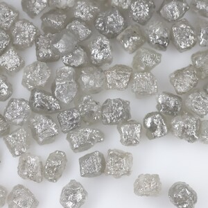 May include: A close-up view of a collection of small, rough, grey diamonds. The diamonds are scattered on a white surface.
