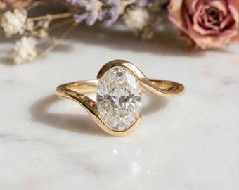 Può includere: Un anello di fidanzamento in oro con un diamante taglio ovale. L'anello presenta un design a spirale, con il diamante incastonato in una lunetta. L'anello è esposto su una superficie di marmo bianco, con fiori sullo sfondo.