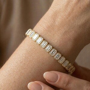 Op de afbeelding: Een gouden armband met een reeks smaragdgeslepen, heldere edelstenen. De armband wordt om een pols gedragen, waarbij de schittering van de stenen wordt benadrukt. De zetting lijkt van goud te zijn, wat de edelstenen versterkt. Een fijn sieraad.