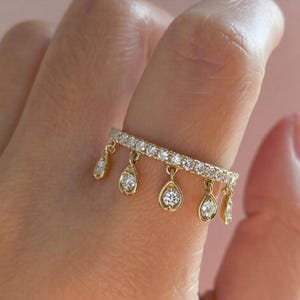 May include: A gold ring with a row of small, clear gemstones and dangling teardrop-shaped pendants, each with a single, clear gemstone. The ring is worn on a finger against a soft pink background.