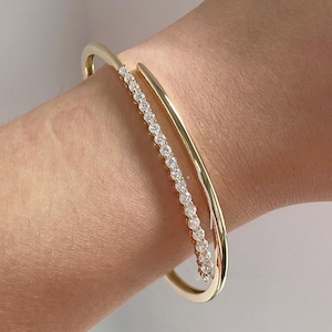 Könnte beinhalten: Ein goldfarbenes Armreif-Armband mit einer Reihe kleiner, funkelnder Steine. Das Armband hat ein schlankes, modernes Design mit einer geschwungenen Form, die das Handgelenk umschließt. Die Steine sind in einer Reihe an einer Seite des Armbands angeordnet.