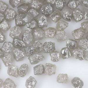 May include: A close-up view of a collection of small, rough, gray diamonds. The diamonds are scattered on a white surface.
