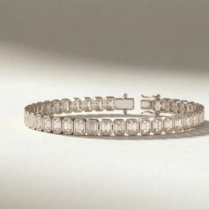 Op de afbeelding: Een zilveren armband met rechthoekige, heldere edelstenen in een bezel-ontwerp. De armband ligt op een witte ondergrond en heeft een sluiting. De edelstenen zijn gelijkmatig verdeeld.