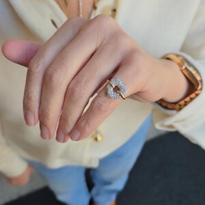 May include: A gold ring featuring a heart-shaped centerpiece encrusted with small, clear, sparkling stones. The band is thin and polished, connecting to the sides of the heart design, which sits prominently on the finger.