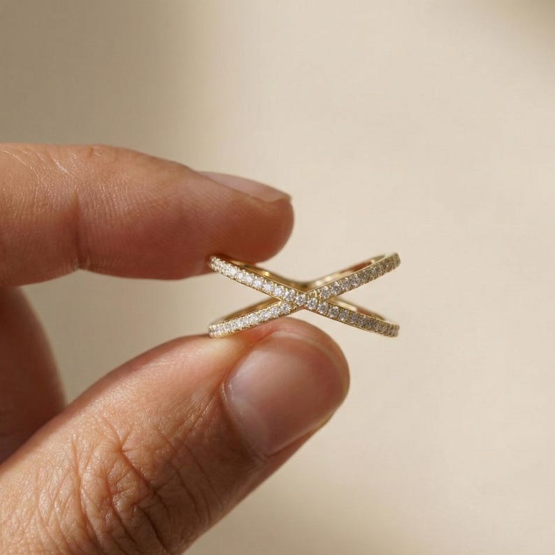 Puede incluir: Un anillo dorado en forma de cruz, incrustado con peque&ntilde;as y brillantes piedras preciosas. El anillo se sostiene entre los dedos sobre un fondo neutro, mostrando su intrincado dise&ntilde;o y su elegante apariencia. El dise&ntilde;o del anillo presenta dos bandas que se cruzan.