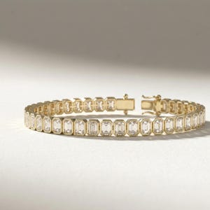 Op de afbeelding: Een gouden armband met een reeks rechthoekige, heldere edelstenen. De armband heeft een gouden sluiting en staat tegen een witte achtergrond. De edelstenen zijn gelijkmatig verdeeld.